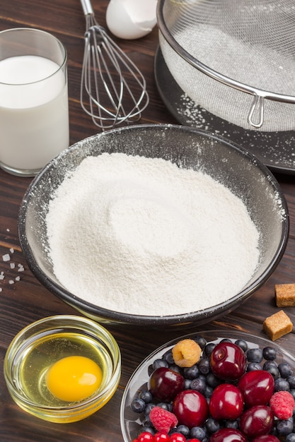 Berries, sifted flour in black plate, cocoa powder. measuring\
cup with flour, glass of milk, broken egg and salt, metal whisk on\
table.