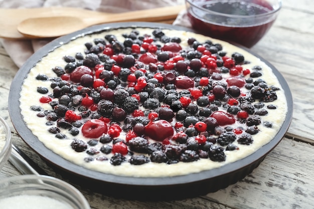 Torta di frutti di bosco cheesecake con mirtillo ribes ciliegia mora con stoviglie e ingredienti sulla tavola di legno.