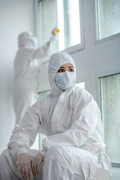 Beschermende maatregelen. Moe blonde medische werknemer in beschermende kleding en medische masker zitten naast het raam, haar collega desinfecterende ruiten