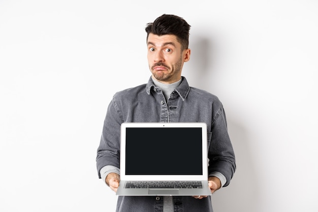 Besluiteloze jongeman pruilend en schouderophalend terwijl hij een leeg laptopscherm laat zien, staande geen idee op een witte achtergrond.