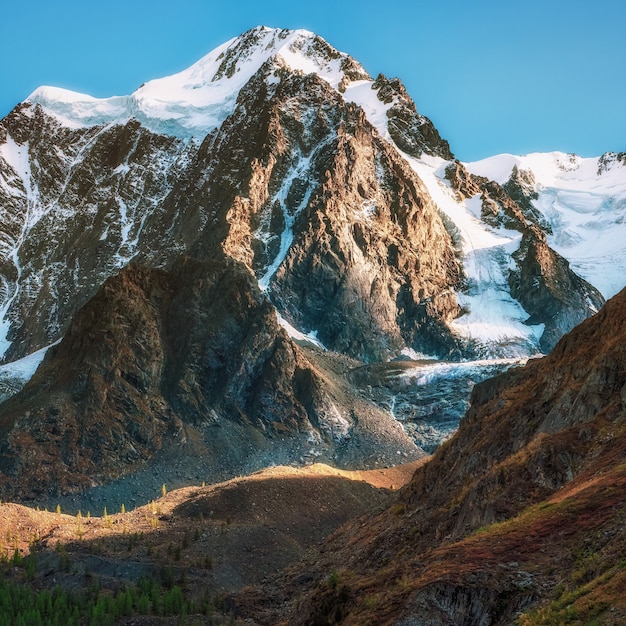 Besneeuwd alpine landschap op grote hoogte met besneeuwde bergtop en scherpe rotsen onder de blauwe hemel.