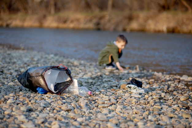 Bespaar milieuconcept, kleine jongen die afval en plastic flessen op het strand verzamelt om in de prullenbak gedumpt te worden