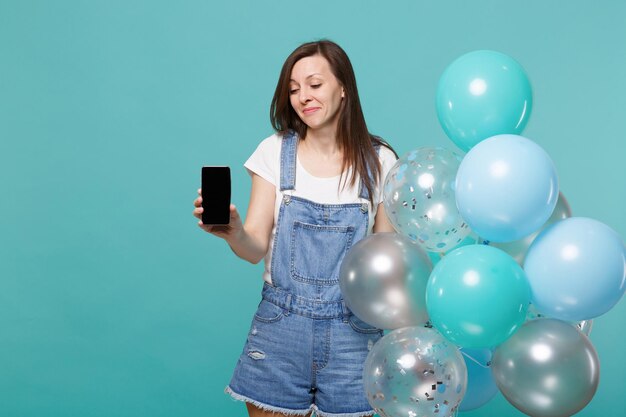 Bespotten van jonge vrouw met mobiele telefoon met leeg leeg scherm, vieren met kleurrijke lucht ballonnen geïsoleerd op blauwe turquoise muur achtergrond. vakantie verjaardagsfeestje, mensen emoties concept.