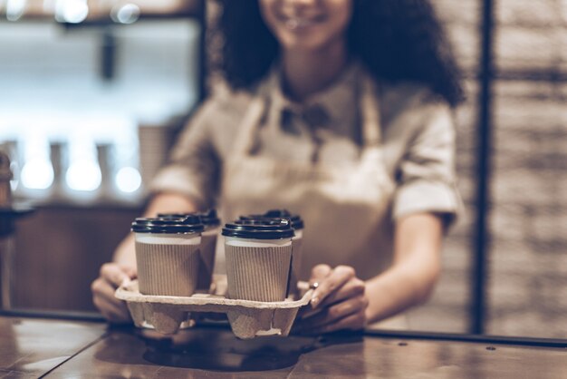 行くのに最適なコーヒー！カフェに立っている間コーヒーカップを保持しているエプロンで若い陽気なアフリカの女性の一部