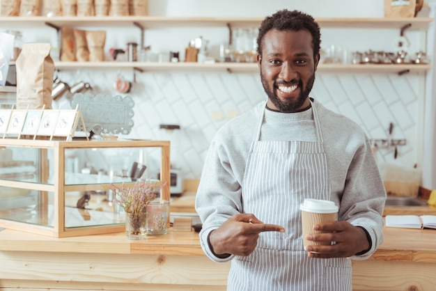 Best coffee. Handsome young male barista in an apron standing in front of a cafe counter and pointing at the cup of coffee in his hands while smiling at the camera