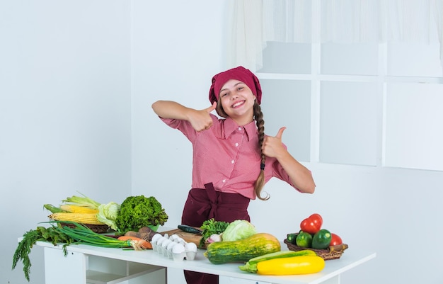 Best day ever full of vitamins kid wear chef uniform and hat teen girl preparing meal fresh raw vegetables time to eat thumb up happy child cooking in kitchen healthy and organic food only