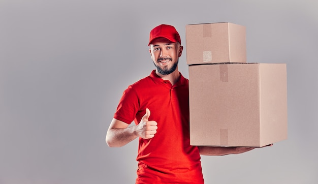 The best delivery service. Cheerful young courier holding a cardboard box and showing his thumb up while standing against gray background