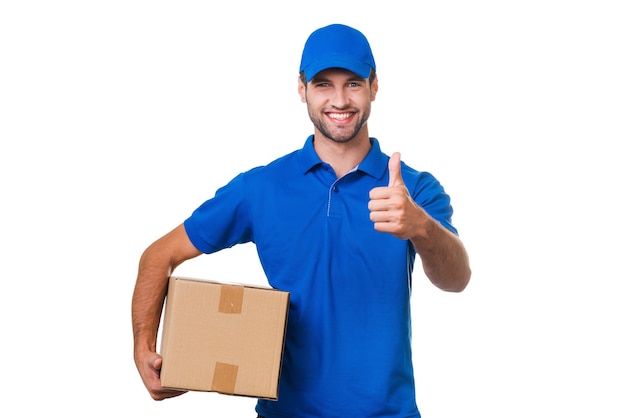 The best delivery service. Cheerful young courier holding a cardboard box and showing his thumb up while standing against white background