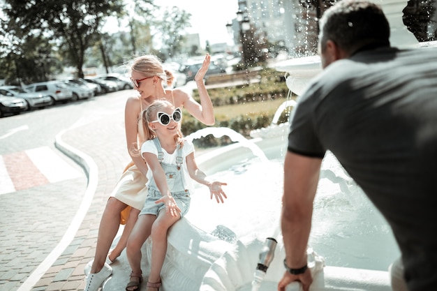Best family. Joyful little girl playing with her parents near beautiful white fountain in the city.
