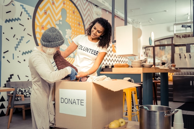 Best fit. Good looking friendly woman smiling while helping an aged woman to find some clothes