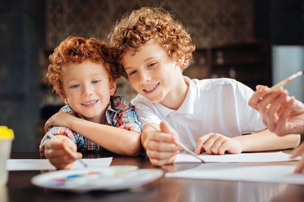 Best friends forever. Portrait of two cheerful curly haired bothers grinning broadly while leaning on each other and enjoying the painting process while sitting at a table.