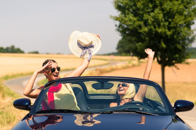 Best friends having summer joyride in convertible car passing a field