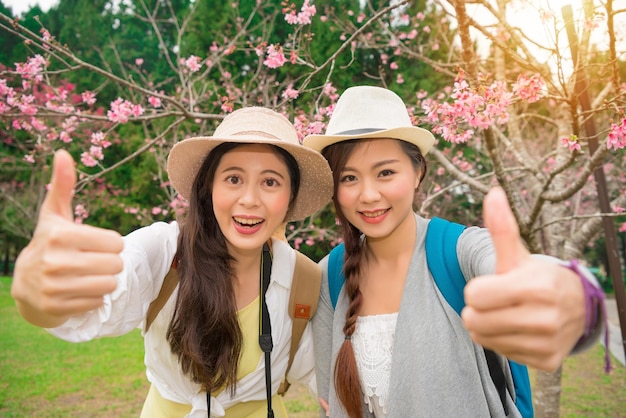 日本の桜公園で2人のスタイリッシュな女の子の身振り手で親指を立てる親友。姉妹旅行で楽しんで幸せな魅力的な若い女性のポートレート、クローズ アップ。