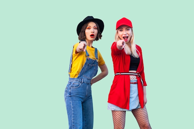 Best friends spending great time together. Two beautiful amazed fashionable hipster girls standing and pointing finger to you, looking at camera. Indoor studio shot, isolated on green background.