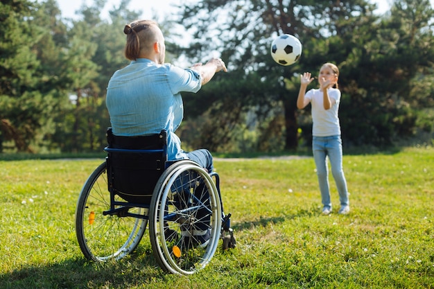 Best friends. Young man with disabilities sitting in a wheelchair and playing volleyball with his adorable little daughter during their walk in the park