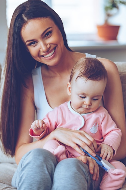 Best mom in the world. Beautiful young woman holding baby girl with toy on her knees and looking at camera with smile while sitting on the couch at home