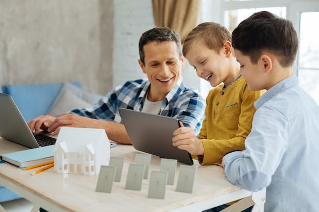 Best pastime. Cheerful pre-teen boys showing their father the video on the tablet while the man taking a break from his work on the laptop