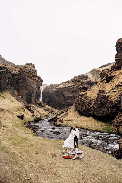 Bestemming IJsland bruiloft, nabij Kvernufoss waterval. Een bruidspaar staat onder een plaid bij een bergrivier. De bruidegom omhelst bruid. Ze bouwden een geïmproviseerde bruiloftstafel met decor en gitaar
