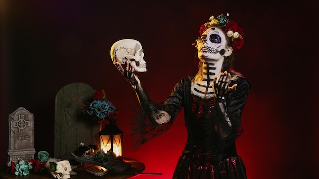 Betoverende dame des doods die schedel over zwarte achtergrond bekijkt, santa muerte-kostuum met bloemenkrans dragend. Zich gedragen als heilige Mexicaanse entiteit op Dios de los Muertos, met festival make-up.