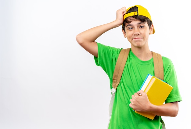 betreurde jonge schooljongen die een rugzak draagt met een pet die boeken vasthoudt en hand op het voorhoofd legt dat op een witte muur wordt geïsoleerd