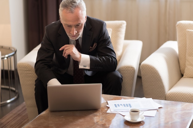 Betrokken aangename bebaarde zakenman die in het hotel zit terwijl hij met de laptop werkt en geconcentreerd is