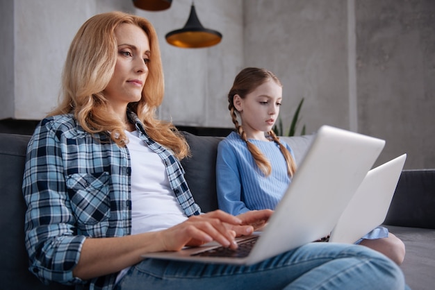 Betrokken charismatische drukke vrouw zittend op de bank en laptops gebruikt met dochter tijdens het surfen op internet en het typen van berichten