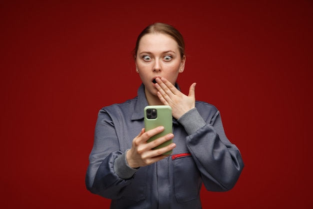 Betrokken jonge blonde vrouwelijke ingenieur die eenvormig bedrijf draagt en naar mobiele telefoon kijkt die hand op mond houdt