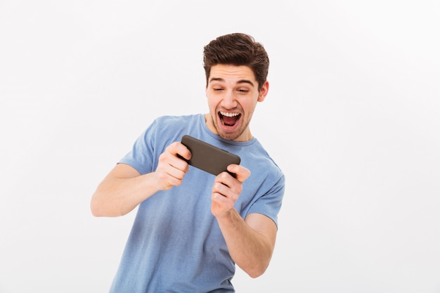 Betrokken man in casual t-shirt spelen van online games op mobiele telefoon, geïsoleerde over witte muur