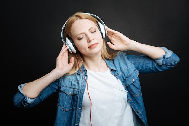 Betrokken vrolijke jonge vrouw die verrukking uitdrukt en glimlacht tijdens het gebruik van een hoofdtelefoon en in de studio staat