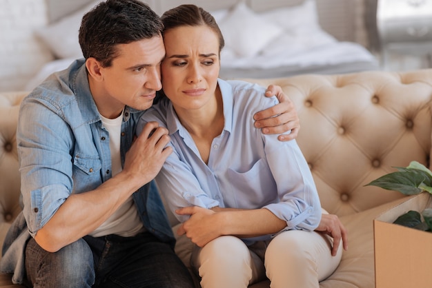 Better. Attentive kind loving man sitting on the sofa and hugging his sad unhappy dismissed wife while trying to encourage her