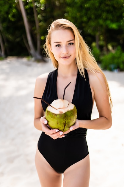 Bella ragazza castana in bikini nero che beve il latte di acqua di cocco vicino all'oceano