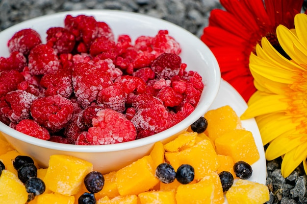 Bevroren fruit, framboos, mango en bosbes in een schaal met een bloem ernaast