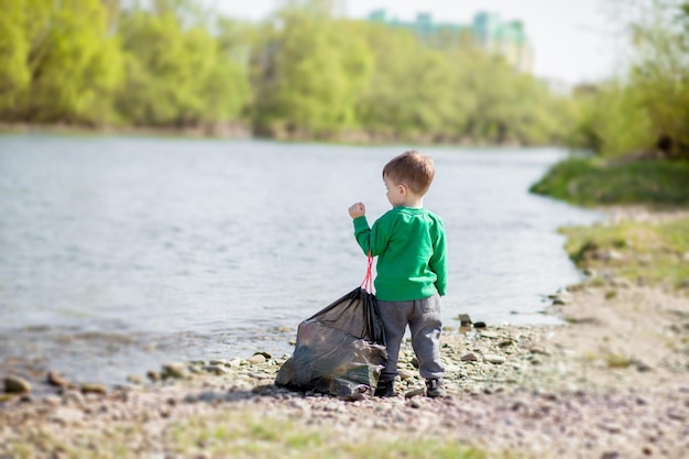 Bewaar het milieuconcept een kleine jongen die afval en plastic flessen op het strand verzamelt om in de prullenbak te dumpen