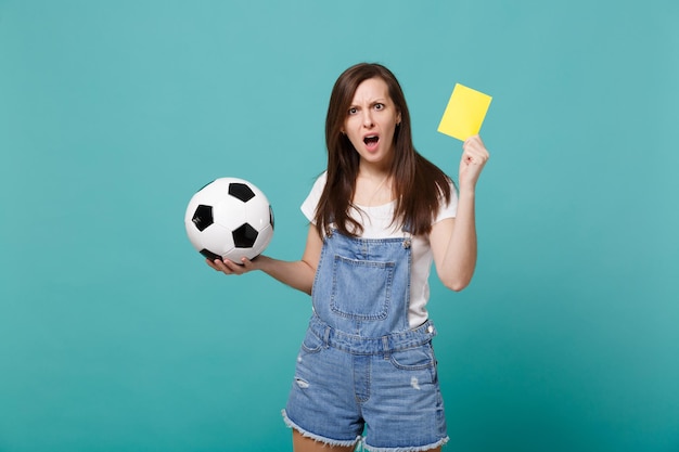 Bewildered girl football fan support team with soccer ball, yellow card, swearing propose player retire from field isolated on blue turquoise background. People emotions, sport family leisure concept.