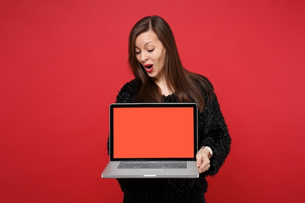 真っ赤な壁の背景に分離された空白の空の画面でラップトップPCコンピューターを探している黒い毛皮のセーターで当惑した若い女性。人々の誠実な感情のライフスタイルの概念。コピースペースをモックアップします。