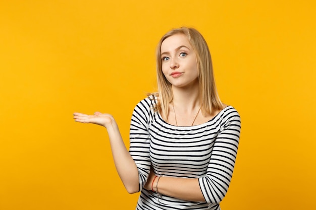 Bewildered young woman in striped clothes looking camera, spreading, pointing hand aside isolated on yellow orange background in studio. People sincere emotions, lifestyle concept. Mock up copy space.