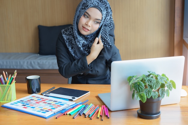 Bezige Aziatische moslim bedrijfsvrouw die op mobiele telefoon in modern bureau spreekt.