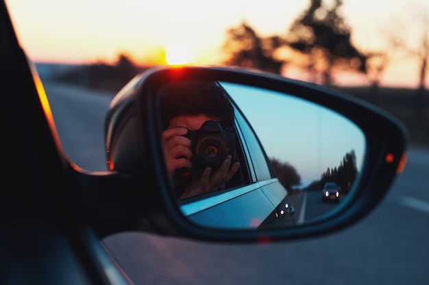 Bezinning in autospiegel van jonge kerel die foto van zonsondergang met digitale camera neemt.