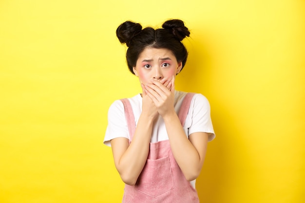 Bezorgd aziatisch tienermeisje die mond bedekken met handen, bezorgd en angstig kijken, bang staan op gele achtergrond met glamoureuze make-up en zomerkleding