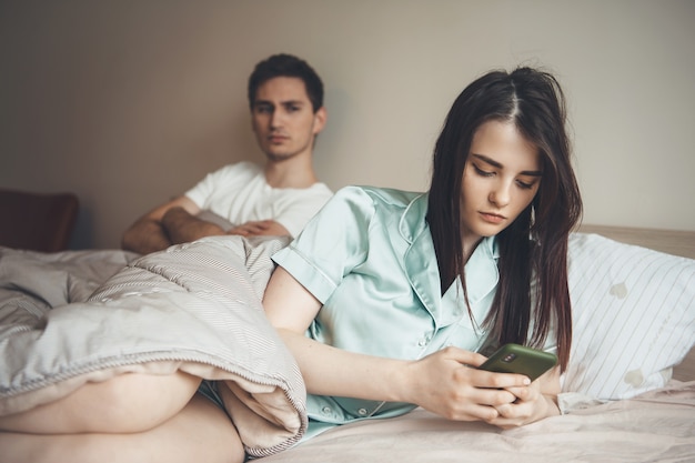 Bezorgd blanke man kijken naar zijn vriendin chatten in bed op mobiel