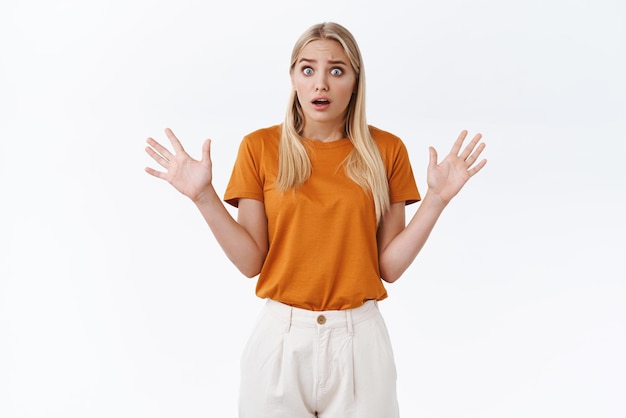 Bezorgd, geschrokken jong blond meisje in oranje t-shirt kijkt bezorgd en geschokt handen omhoog zijwaarts open mond en hijgend bezorgd, twijfelachtig en onzeker witte achtergrond