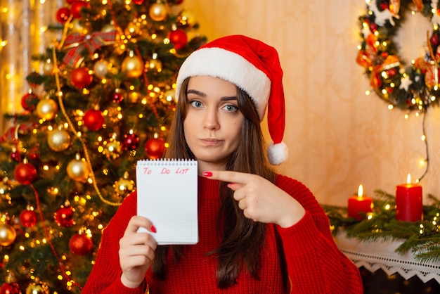 Bezorgd jong meisje in kerstmuts en rode trui met notitieboekje en wijzend op takenlijstzin Veel dingen om te doen voor de kerstvakantie