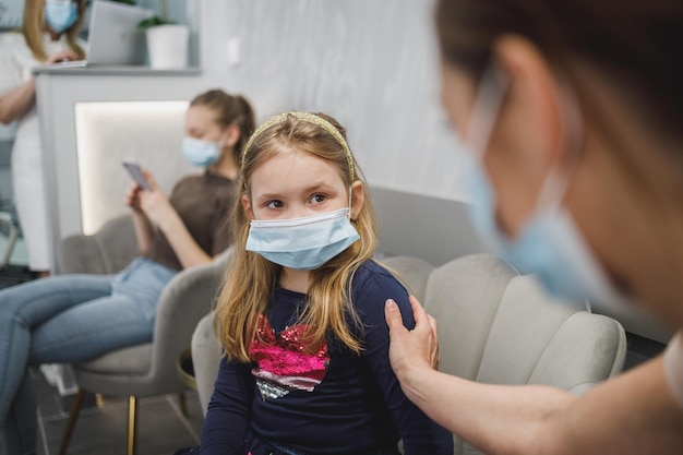 Bezorgd klein meisje en haar moeder met gezichtsmasker in wachtkamer op het kantoor van de tandarts.