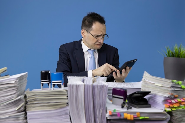 Bezorgd zakenman rekent op calculator zittend aan tafel met stapels papieren in kantoor