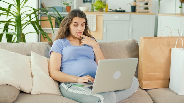 Bezorgd zwangere vrouw online kopen met behulp van Laptop en creditcard in de gezellige woonkamer thuis met moderne interieur Financiën Online winkelen fouten en mislukkingen