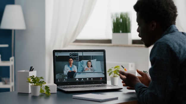 Bezorgde Afro-Amerikaanse vader voor klein meisje in ziekenhuisafdeling, pratend met arts op videoconferentiegesprek met webcam-app op afstand scherm voor familieverbinding. Huisarts consult via internet