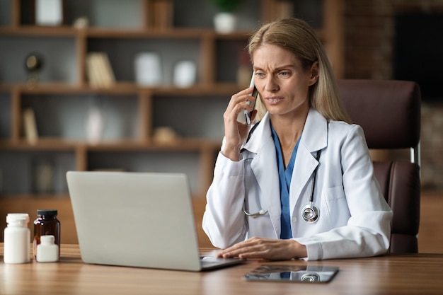 Bezorgde dokter vrouw met behulp van laptop en praten over mobiele telefoon op de werkplek