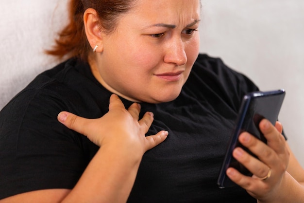 Bezorgde grote roodharige vrouw in zwart t-shirt die slecht nieuws bekijkt op de smartphone die ze vasthoudt in haar...