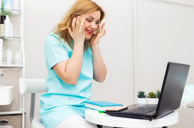 Bezorgde jonge vrouwelijke blanke arts in blauw medisch uniform zit aan bureau in ziekenhuis werk op laptop, arts adviseert en laat zien hoe gezichtsmassage te doen.