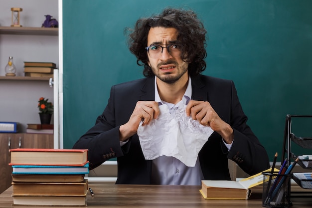 Bezorgde mannelijke leraar die een bril draagt, scheurt het papier omhoog terwijl hij aan tafel zit met schoolgereedschap in de klas